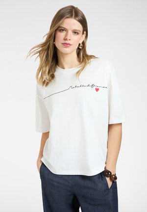 Ung kvinde med blondt hår iført hvid oversized T-shirt med teksten "Make the difference" og et rødt hjerte, kombineret med mørke bukser.