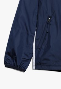 Námořnická modrá větrovka, lehký materiál, elastické manžety, zipová postranní kapsa, bílý perforovaný detail podšívky.