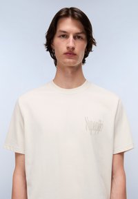 Cream-colored cotton t-shirt features a crew neck and short sleeves, with a subtle logo printed on the left chest in a slightly darker shade.