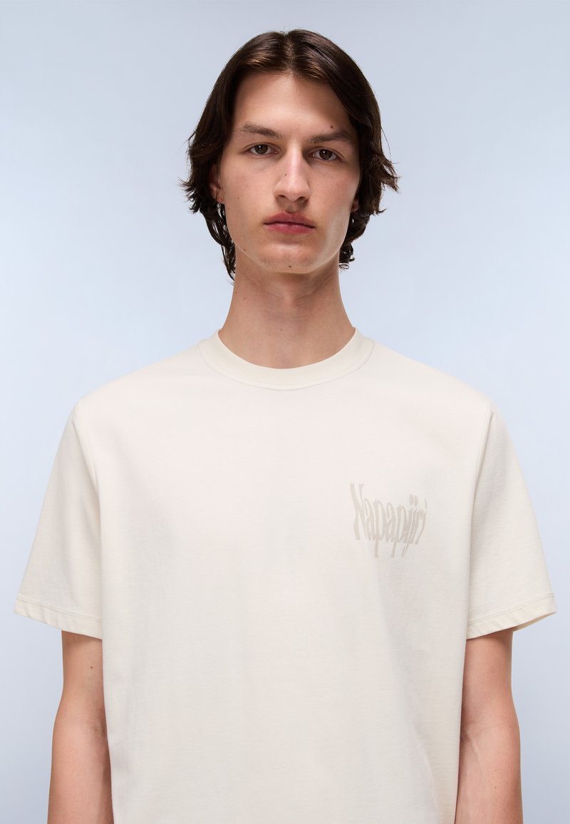 T-shirt en coton crème à col rond et manches courtes, avec un logo subtil imprimé sur la poitrine gauche dans une teinte légèrement plus foncée.