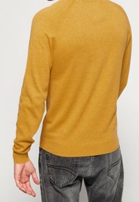 Maglione giallo a maglia con un design aderente, polsini e orlo a coste, visto da dietro. Abbinato a jeans grigio scuro con tasche posteriori.