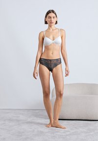Reggiseno bianco in tessuto liscio, abbinato a mutandine nere in pizzo che mostrano intricati motivi floreali. La modella è in piedi su un tappeto grigio chiaro.