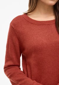 Kvinne iført en rustfarget strikkegenser med ribbestrikket rund hals og lange ermer, vist fra skuldrene til midten av torso.