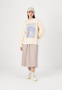 Kremfarget oversized hettegenser med blå grafikk, kombinert med en beige midiskjørt. Modellen har på seg en lys caps og hvite sneakers.