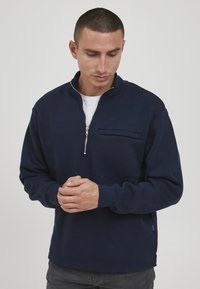 Mann mit kurzen Haaren, der einen marineblauen Quarter-Zip-Pullover und dunkle Hosen trägt, nach unten schaut mit vor ihm gefalteten Händen.