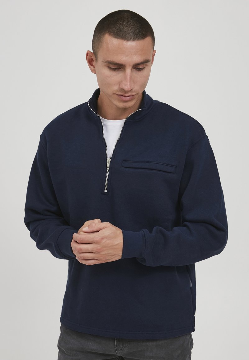 Mann mit kurzen Haaren, der einen marineblauen Quarter-Zip-Pullover und dunkle Hosen trägt, nach unten schaut mit vor ihm gefalteten Händen.