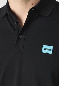 Svart polo skjorte med teksturert stoff, med en ribbestrikket krage, to knapper og en lys blå rektangulær "HUGO" logo brodert på brystet.