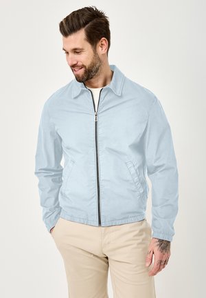 Man with beard wearing light blue zip-up jacket and beige pants, looking down and smiling, hands in pockets.