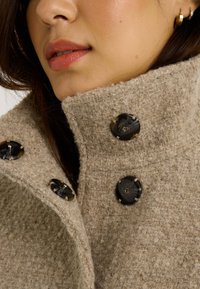 Acercamiento a la parte inferior de la cara de una mujer que lleva un abrigo de textura beige con cuello alto, botones oscuros grandes y un pequeño pendiente de aro dorado.