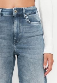 Jeans in denim azzurro chiaro con vita alta, con chiusura frontale a bottoni, tasche laterali e una texture leggermente strappata.