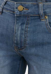 Jeans de mezclilla en azul medio con costuras naranjas. Presentan un cierre de botón, cremallera y bolsillos frontales en ángulo. Textura de tela suave.