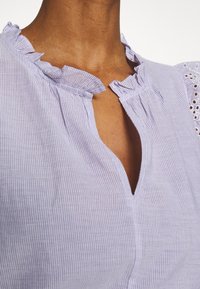 Vaaleanliila raidallinen pusero, jossa on röyhelöinen pääntie ja avainreikäaukko; hihat koristeltu herkillä pitsinauhoilla. Puuvillasekoitekangas.