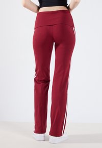 Pantalons de sport bordeaux avec une taille haute, des rayures blanches sur les côtés et une texture lisse. Ils présentent une coupe décontractée et un design de jambe fuselée.
