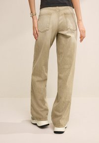 Personne portant un pantalon beige ample, un haut noir, des baskets blanches et une montre, se tenant avec les mains détendues sur les côtés devant un fond uni.