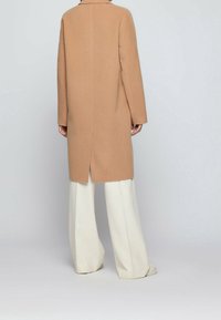 Cappotto di lana cammello con silhouette dritta, maniche lunghe e uno spacco sul retro. Abbinato a pantaloni bianchi a gamba larga e scarpe casual.