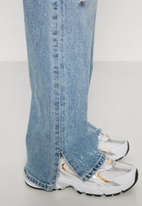 Světle modré džínové kalhoty s třepeným lemem a bočními rozparky, doplněné bílými a stříbrnými sportovními botami s oranžovými akcenty.