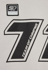Gaiši pelēka auduma izstrādājums ar lielu melnu numuru "71" un tekstu "TEAM OF CHM PNS." Ietver kvadrātveida melnu etiķeti ar baltu tekstu un logo.