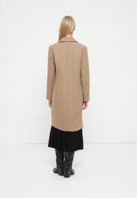 Cappotto lungo da donna color cammello con colletto strutturato e vestibilità dritta, abbinato a una gonna nera plissettata e stivali neri al ginocchio.