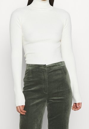 Femme portant un col roulé côtelé blanc ajusté et un pantalon en velours vert foncé taille haute, debout devant un fond uni.