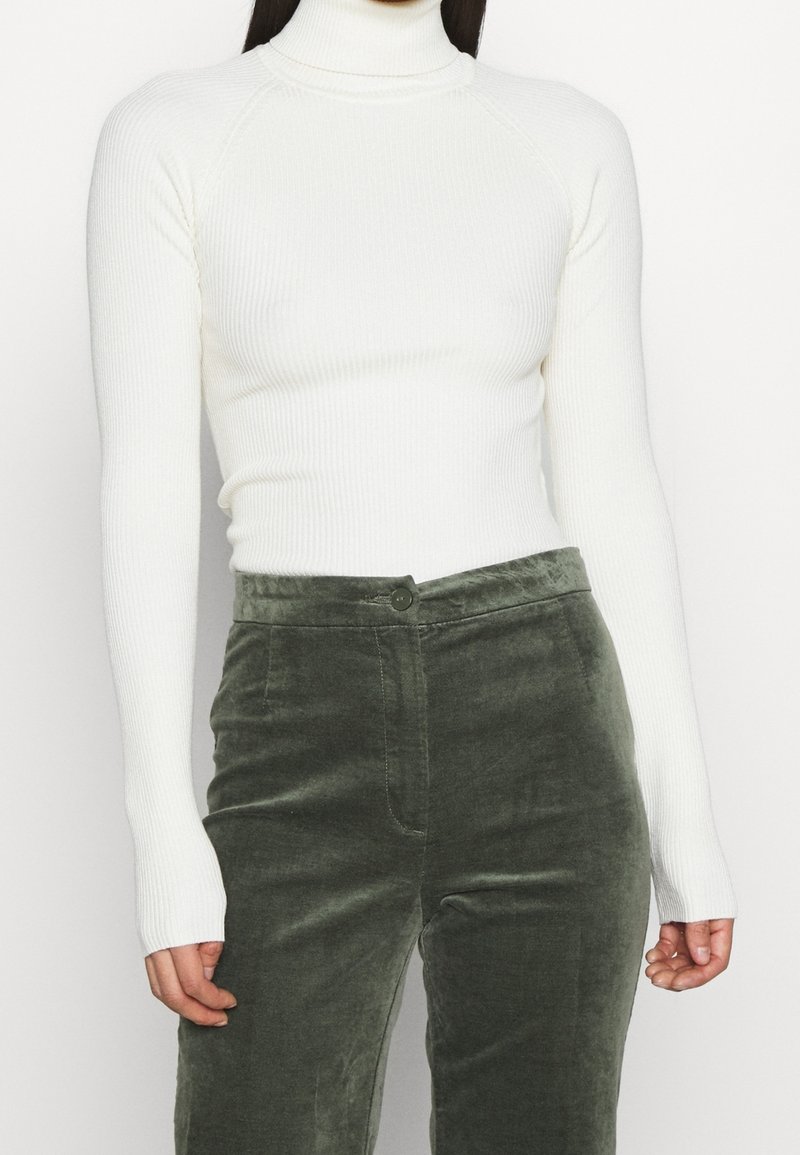 Femme portant un col roulé côtelé blanc ajusté et un pantalon en velours vert foncé taille haute, debout devant un fond uni.