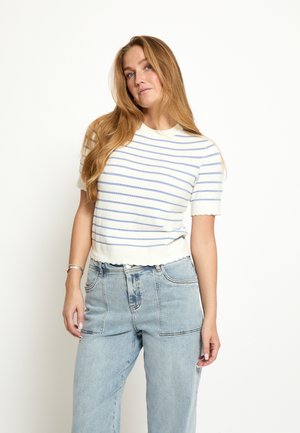 Frau mit langen blonden Haaren, die ein weißes kurzärmeliges Oberteil mit blauen Streifen und hellblaue hoch taillierte Jeans trägt, vor einem weißen Hintergrund.