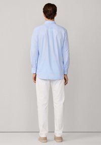 Camisa de manga larga azul claro con cuello abotonado y pliegue en la espalda, combinada con pantalones blancos y zapatos color cognac. Diseño simple y limpio.