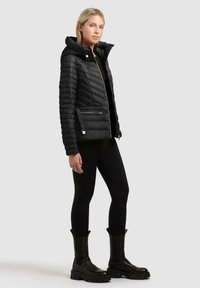 Donna in piedi che indossa una giacca nera trapuntata con cappuccio, leggings neri e stivali neri robusti su uno sfondo grigio uniforme.