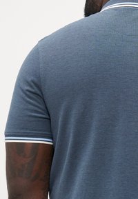 Man in een blauwe polo met een witte en blauwe gestreepte kraag en mouwboorden, met een getatoeëerde bovenarm en een donkere huid.