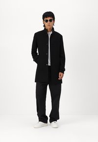 Abrigo negro de botones con corte recto y cuello alto. Lleva una camisa blanca y pantalones oscuros. Presenta bolsillos frontales y gafas de sol.