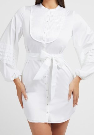 Femme portant une robe blanche boutonnée avec des manches bouffantes, un plastron plissé à l'avant, et une ceinture nouée à la taille, les mains posées sur les hanches.