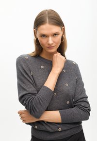 Grauer Strickpullover mit runden goldenen Akzenten, im lässigen Design. Hat lange Ärmel und gerippte Bündchen, glatte Textur.
