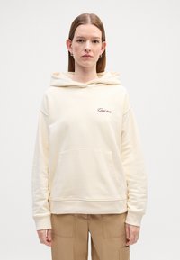 Hoodie couleur crème fabriqué en tissu doux, présentant une poche avant, une capuche à cordon de serrage et un logo texte subtil en rouge foncé sur la poitrine.