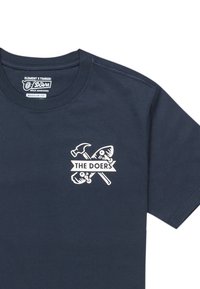 Tmavě modré tričko v běžném střihu, s bílou grafikou rukou držících nářadí a textem "THE DOERS" na levé části hrudi.