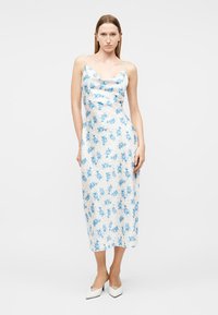 Γυναίκα που φοράει λευκό midi φόρεμα με μπλε λουλουδένιο μοτίβο και λευκές μυτερές γόβες, στέκεται μπροστά σε απλό λευκό φόντο.