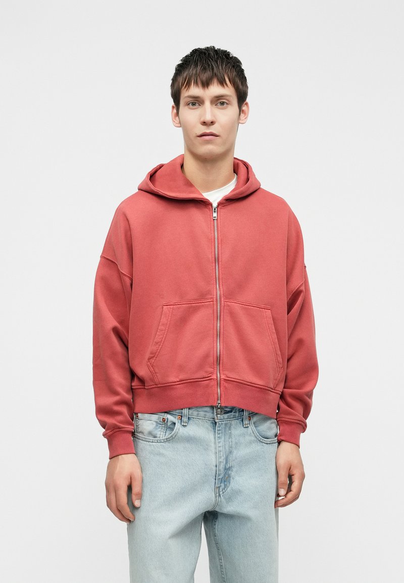 Κόκκινη ζακέτα με φερμουάρ, διαθέτει μπροστινή τσέπη και κουκούλα με κορδόνι, φτιαγμένη από μαλακό ύφασμα. Συνδυάζεται με ανοιχτό γαλάζιο τζιν παντελόνι.