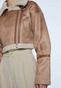 Bruine cropped jas van imitatiesuède met schapenvachtkraag, ritssluiting en bruine lederen accenten; gecombineerd met lichtbeige, hoog uitgesneden broek.