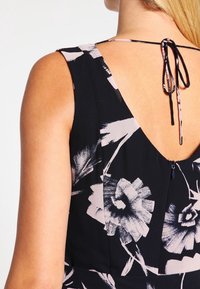 Robe fleurie noire avec un dos en V profond, fermeture par lien délicat, et un tissu doux et texturé avec de grands motifs floraux clairs.