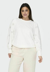 Felpa bianca con maniche a sbuffo e dettagli a intaglio floreali. Il tessuto sembra morbido, abbinato a pantaloni chiari e texturizzati.