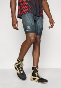 Ανδρικά εφαρμοστά σορτς σε σκούρο τιρκουάζ με μοτίβα που μοιάζουν με φύλλα και χρυσές λεπτομέρειες· λογότυπο στο αριστερό μηρό. Φορεμένα με μαύρα και καφέ αθλητικά παπούτσια ψηλού τύπου.