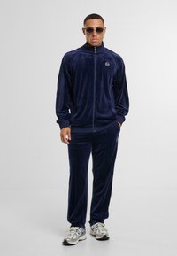 Mężczyzna ubrany w granatowy dres z weluru z zamkiem błyskawicznym oraz dopasowane spodnie, w srebrnych sneakersach i okrągłych okularach przeciwsłonecznych, stoi na białym tle.