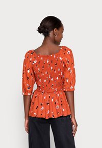 Blusa vermelha com padrão, featuring mangas bufantes, corpete franzido e bainha rodada. Os padrões incluem desenhos florais em branco e azul.
