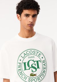 Camiseta de algodón blanca con un logo circular verde que incluye el texto "LACOSTE PARIS FRENCH SPORTSWEAR" y un detalle gráfico de un cocodrilo.