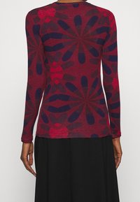 Osoba v dlouhém rukávu vínového topu s velkými tmavě modrými a červenými květinovými vzory, v kombinaci s černou sukní.