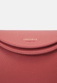 Różowa skórzana torebka o teksturowanej powierzchni, zaokrąglone zapięcie na klapę oraz złote wytłoczone logo marki z przodu.