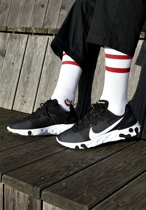 Ben iført sorte bukser, hvide sokker med røde striber og sort-hvide Nike React-sneakers, der hviler på trætrin udendørs.