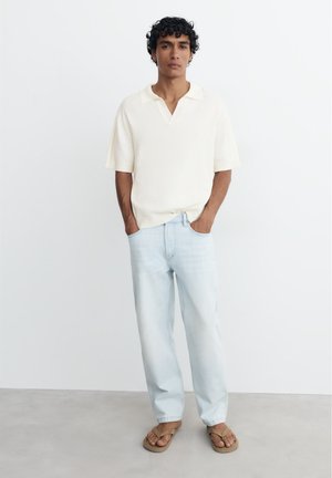 Mand med krøllet hår iført en hvid kortærmet skjorte, lyseblå jeans og beige sandaler står med hænderne i lommerne.