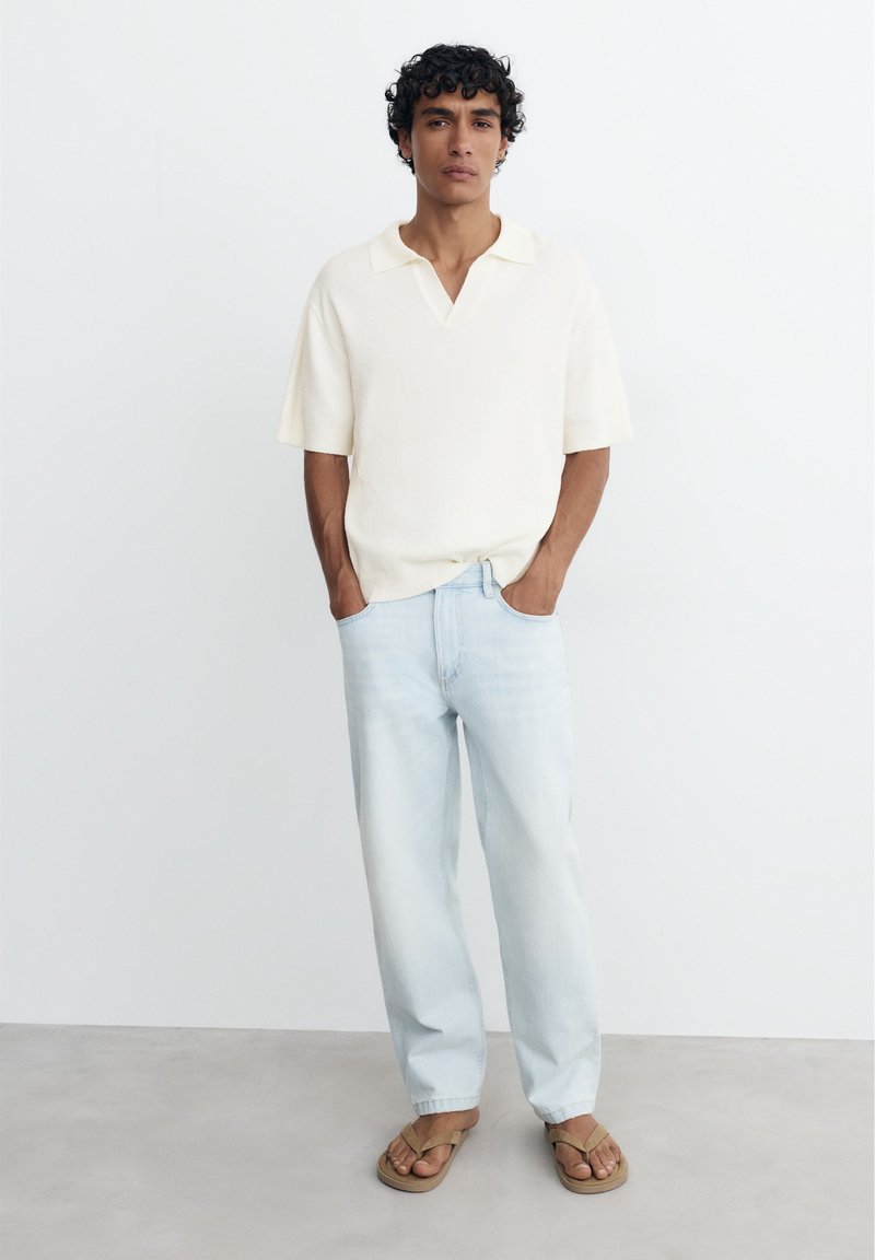 Homme aux cheveux bouclés portant une chemise blanche à manches courtes, un jean bleu clair et des sandales beiges, debout les mains dans les poches.
