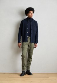 Dunkelblauer Knopf-Cardigan mit strukturiertem Strick, kombiniert mit olivgrünen Cargo-Hosen mit elastischen Bündchen. Schwarze Knöchelstiefel runden das Outfit ab.
