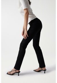 Pantalones de mezclilla negros con un diseño ajustado, ligeramente elásticos y un acabado limpio. Combinados con sandalias de tacón negro minimalistas y una blusa blanca con canalé.