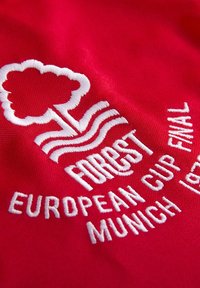 Rød stof med hvidt broderet tekst og grafik. Designet indeholder et træ og bølger, med "FOREST," "EUROPEAN CUP FINAL," og "MUNICH 1980."